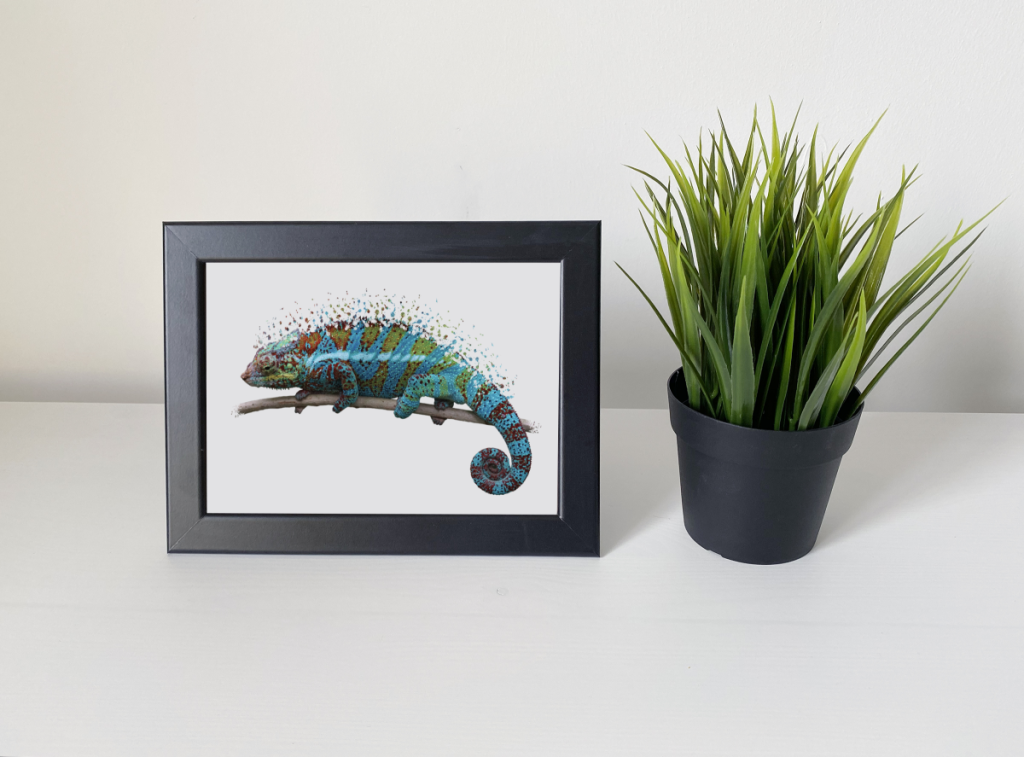 A photo of a lizard wall art in a black frame. There is a plant to the side of it in a black pot.